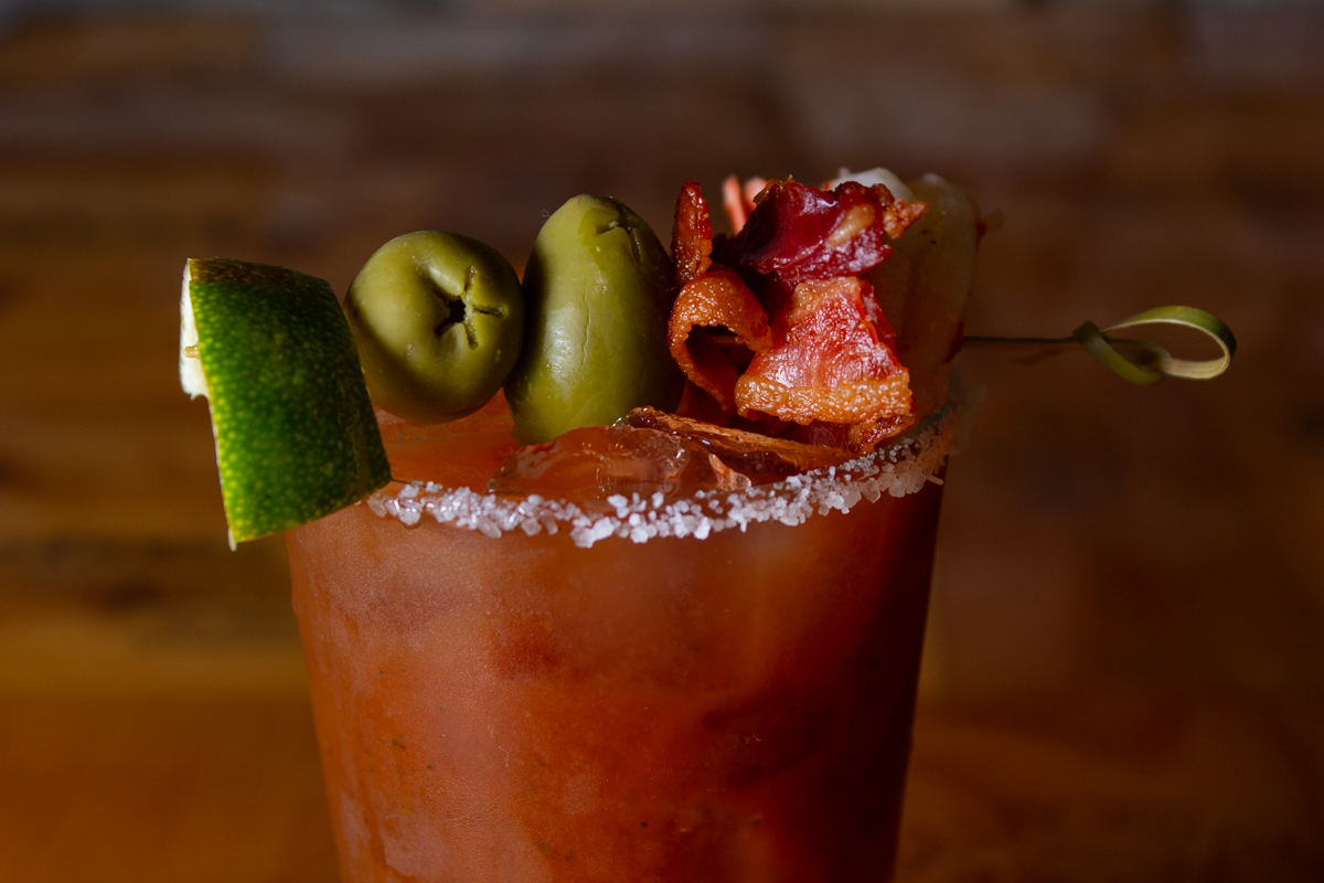 Stonewater_Bloody_Mary_20220830 - 002comp Example of Food Photography for DoorDash, Uber Eats, Grub Hub, and any food delivery service app. Food Photo is of a beverage prepped by a restaraunt. It shows the drink from the top half of the glass shot at a 45 degree angle so you can still see the drink, but the image focuses on the garnish on top which is a screwer with green apple, olives, and baco