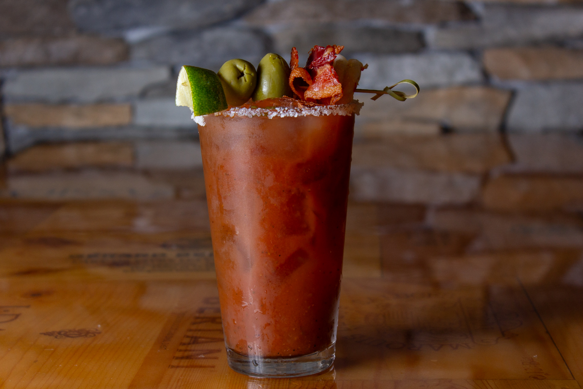 Stonewater_Bloody_mary_20220830 - 001comp Example of Food Photography for DoorDash, Uber Eats, Grub Hub, and any food delivery service app. Food Photo is of a drink prepped by the restaraunt. The image is a profile shot showing the side of the brown drink. The drink has a screwer on top with olives, bacon, and green apple. The rim of the glass is lined with sugar salt. The backgorund of the image is a stone wall.