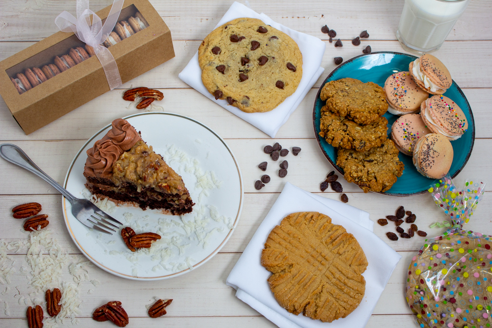 20210531_Letem_Eat_Kake_Bakery_Hero Shot Example of Food Photography for DoorDash, Uber Eats, Grub Hub, and any food delivery service app. Food Photo is of baking goods. There is a piece of chocolate cake, cookies on a platetr, a couple large cookies, and an example of the companies food packaging. There are chocoalte chips sprinkled around for decoration. The background is a light color wood. This is what most fod delivery services call a "Hero Shot" and is used as a banner on a website or on ethe top of the restaraunt portal on the apps.
