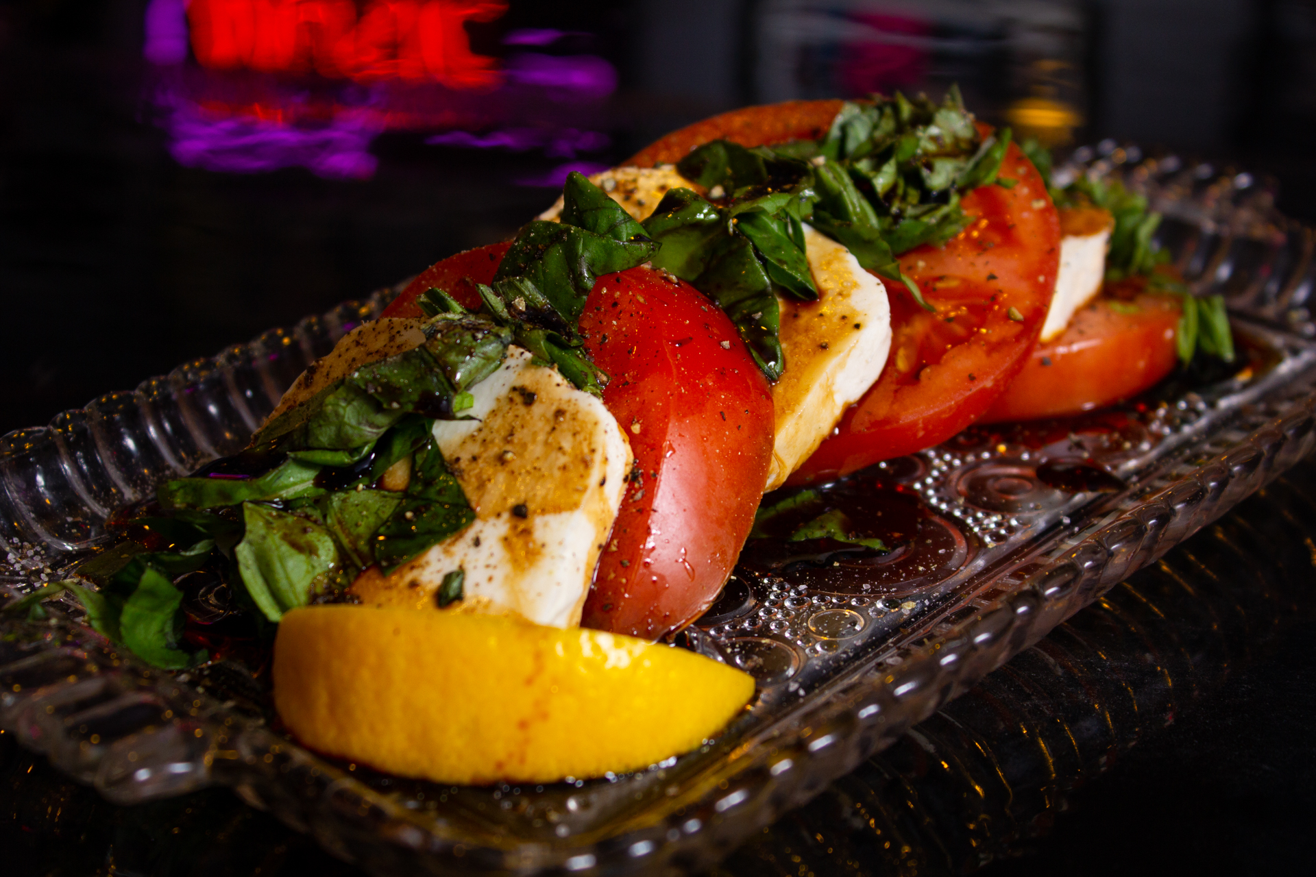 20220131_Rock_Pizza_Subs_(S_Missouri_Ave)_The Caprese_Appetizer Example of Food Photography for DoorDash, Uber Eats, Grub Hub, and any food delivery service app. Food Photo is of a tomato based appetizer garnished with oranges and other greenery, sitting on a glass decorative platter. The background surface is shiny black and is reflecting a neon logo for flare.