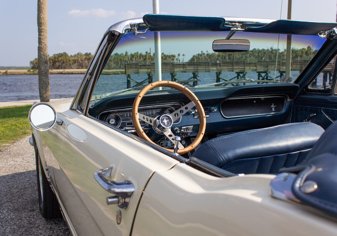 1965_Ford_Mustang_Convertible_20230630 (130)comp Example of Automotive Photography. Car Photography and Videography for Bring-a-Trailer, Auto Hunter, Barn Finder, Auto Trader, and any car and automobile sales sites. Car Photo is of a 1965 Ford Mustang convertible. It is cream colored. The shot is from over the shoulder of the driver with the top down. We are looking at the stearing wheel and the water is in the background of the dashboard. The convertible top is open and we see a nice view of the Gulf.
