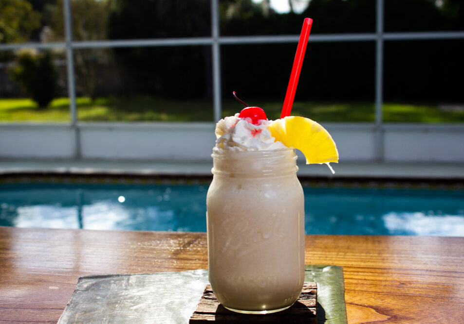 20211109_El_Tropezon_Pina_Colada Example of Food Photography for DoorDash, Uber Eats, Grub Hub, and any food delivery service app. Food Photo is of a Pina Colada, so this is an example of Drink Photography. the photo is taken on a pool deck outside, from the side of the drink. We see the pool in the background. The drink has a pineapple wedge and cherry at the top with a red straw.