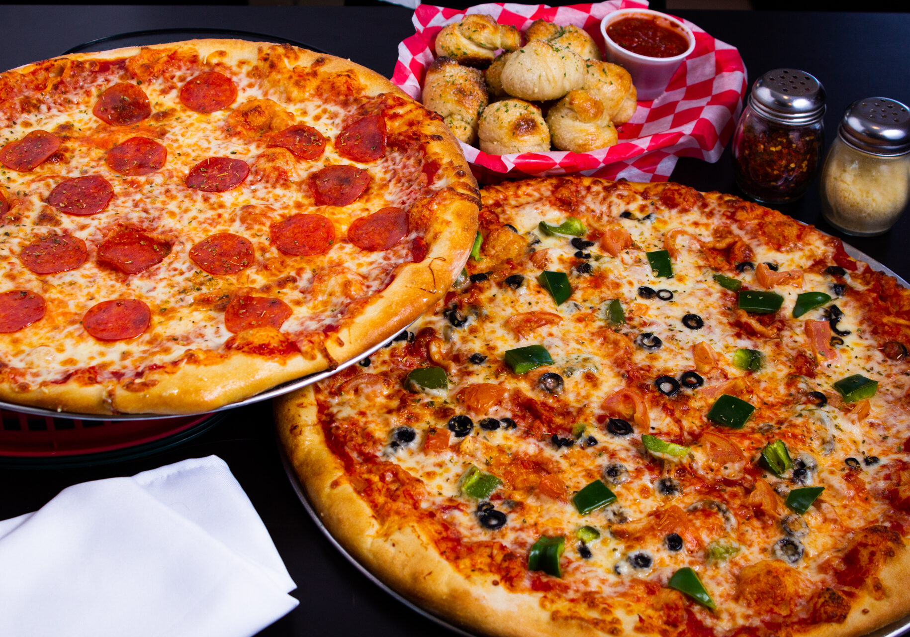 20220131_Rock_Pizza_Subs_(S_Missouri_Ave)_Pizzas Example of Food Photography for DoorDash, Uber Eats, Grub Hub, and any food delivery service app. Food Photo is of a group of pizzas. Has great colors and professionally edited photo. There is a pepperoni pizza, a supreme pizza, and garlic knots. Photo is taken over a dark black granite reflective surface to showcase the food.