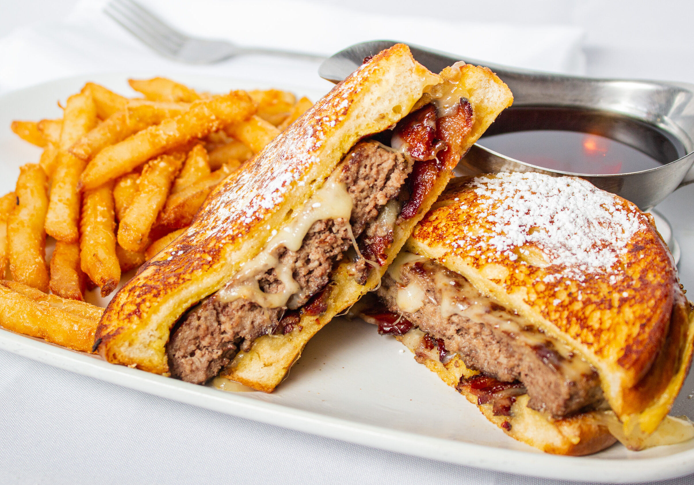 Foodys Diner and Pizza House_French Toast Burger_0009 Example of Food Photography for DoorDash, Uber Eats, Grub Hub, and any food delivery service app. Food Photo is of a hamburger on french toast with syrup on the side. We see the syrup in a syrup gravy boat and there is also a side of fries. The photos is taken over a white background to showcase the food item.